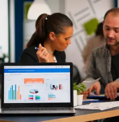 A laptop displaying colorful data visualizations sits on a table as two people engage in discussion with documents and a small plant.