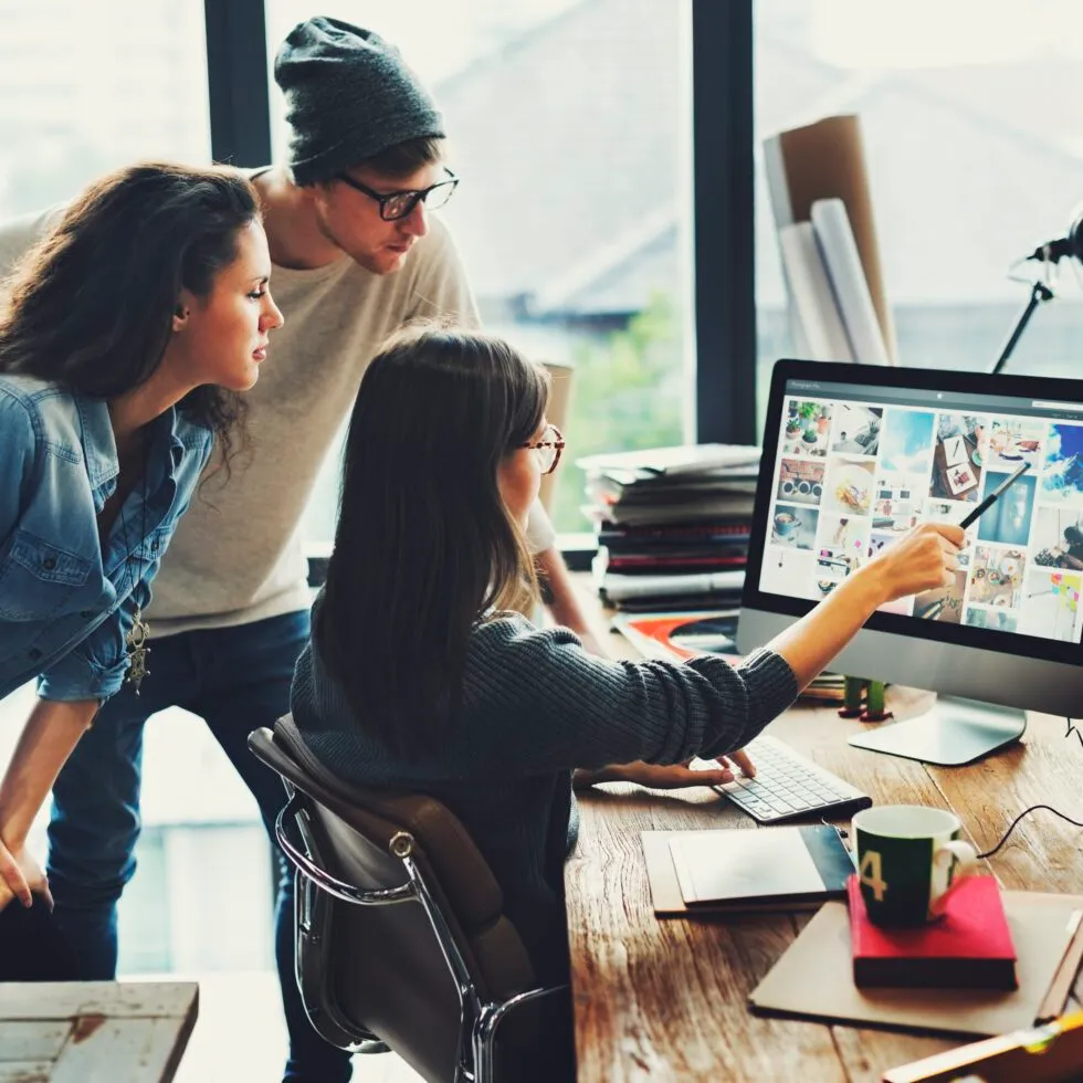 A woman in a sweater points at a computer screen while discussing design ideas with two others in a bright office space.