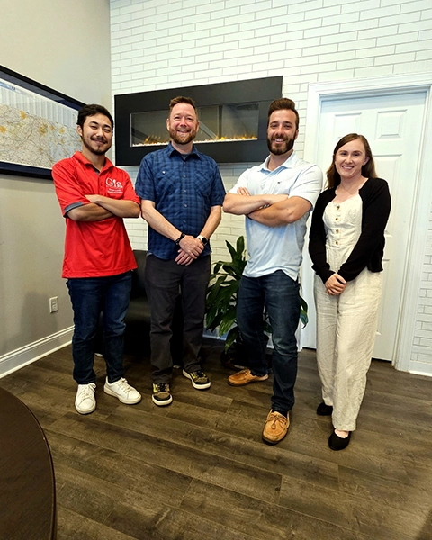 Four individuals stand confidently in an office space, with a plant and modern decor in the background.